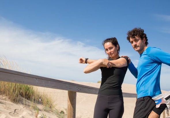 A man and woman in activewear stretch on a sunny Portugal beach boardwalk, promoting fitness and outdoor activities.